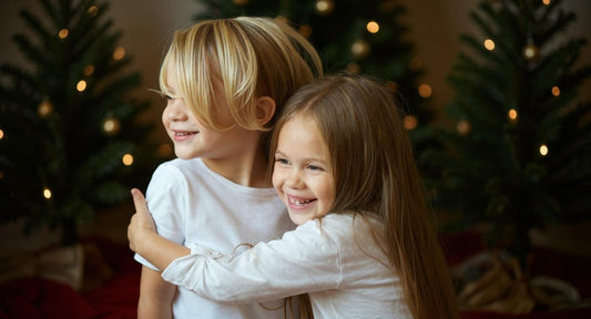 Zwei blonde, strahlende Kinder (Mädchen und Junge) vor einem geschmückten Weihnachtsbaum, die sich auf das Auspacken ihrer Geschenke freuen.