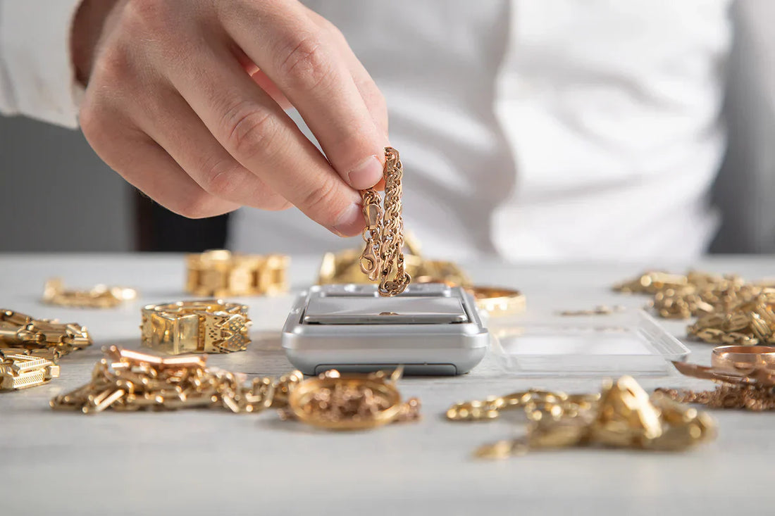 Eine Hand hält Gold-Schmuck bereit, um ihn auf die digitale Goldwaage zu legen. Um die Waage herum liegen weitere Ketten und Ringe aus Gold zur Gewichtsprüfung bereit.