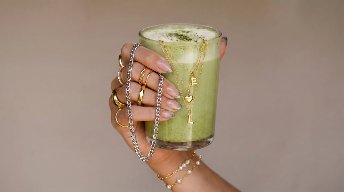 Eine Hand mit mehreren goldenen Ringen, Armbändern und einer silberfarbenen Kette hält ein Glas mit grünem Matcha-Latte. Am Glas hängen drei filigrane goldene Ketten mit Buchstaben- und Herzanhängern.